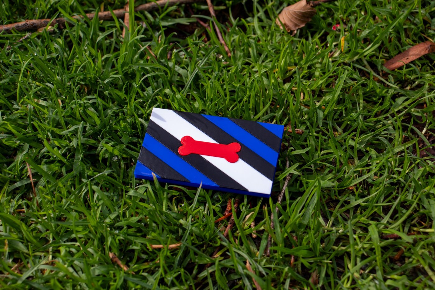 Pup pride flag magnet with blue, black and white stripes and a red bone, lying on grass
